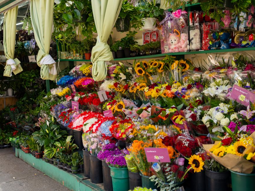 A vibrant floral display outside a florist shop, featuring an assortment of fresh bouquets arranged on tiered shelves and in buckets. Bright sunflowers with large yellow petals and dark centers are pr