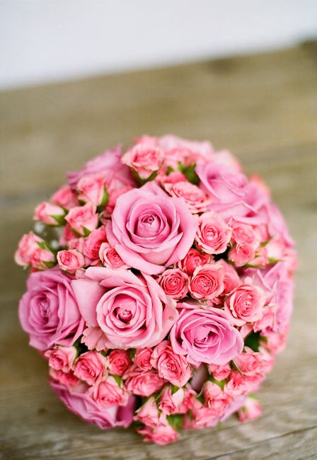 A floral arrangement featuring a dense cluster of pink roses, with some smaller roses and buds interspersed, all arranged in a rounded bouquet. The flowers exhibit fresh, velvety petals with subtle va