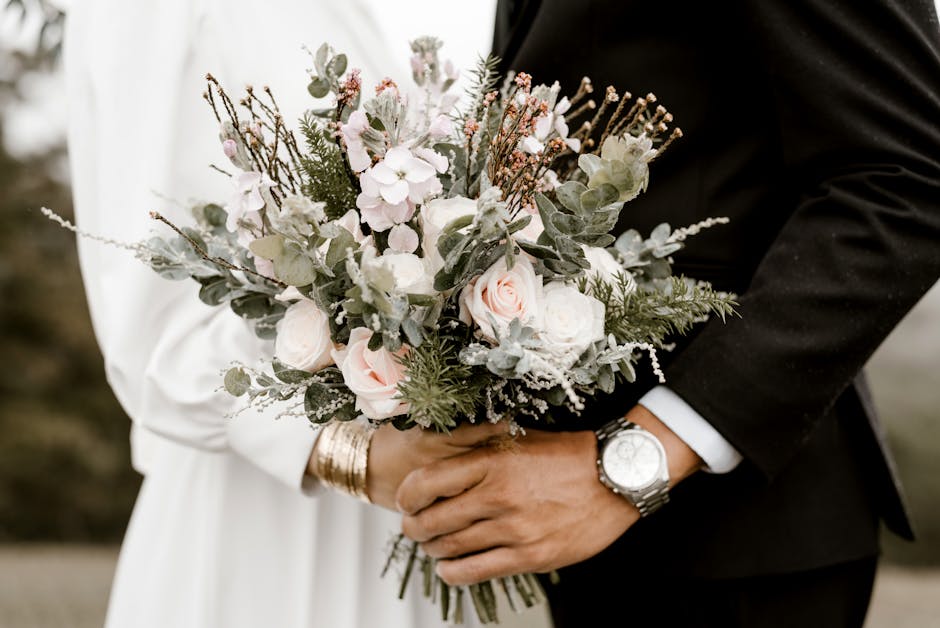 A close-up view of a bridal bouquet held by a bride and groom. The bouquet features soft pink roses, white roses, and pale pink hydrangeas, accented with green and silvery eucalyptus leaves and sprigs