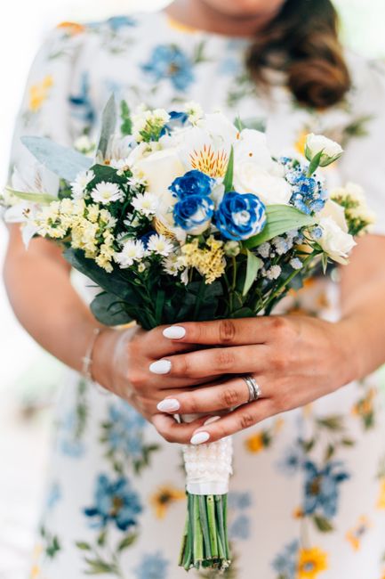 A person holding a fresh floral bouquet featuring white roses, blue carnations, small white daisies, and yellow accents, with eucalyptus leaves and other greenery. The bouquet is wrapped with white ri