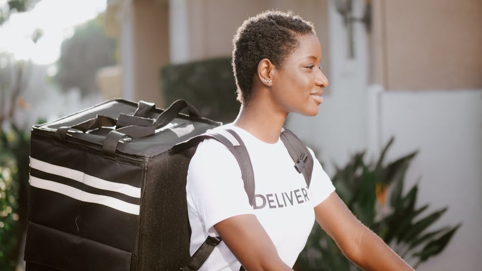A young woman with short, curly hair and medium skin tone, dressed in a white T-shirt with the word 'DELIVER' on it, is holding a bicycle. She is smiling softly and looking ahead as she prepares for a
