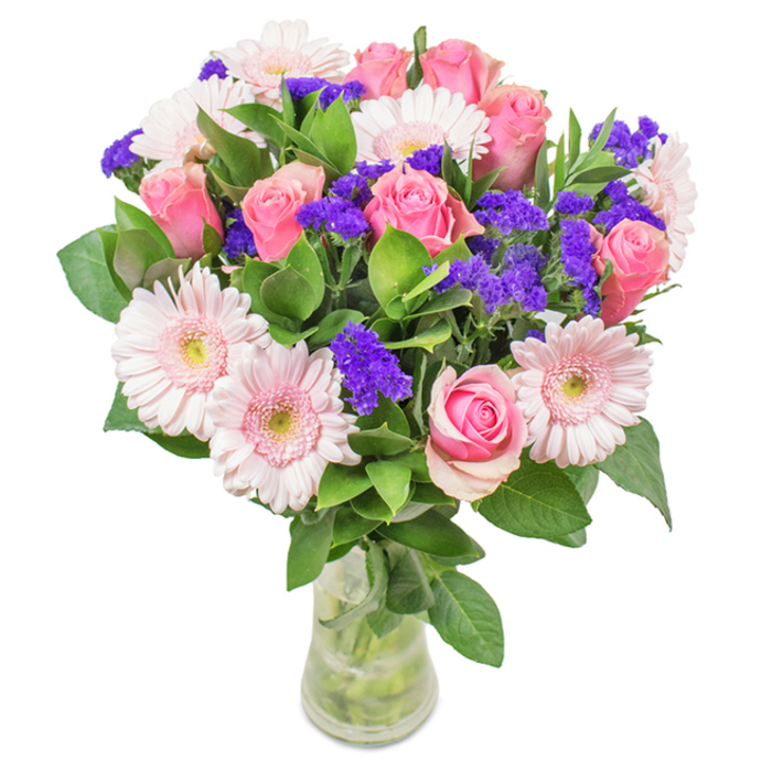 A luminous floral arrangement stands in a clear glass vase, set against a crisp, white background that evokes the clean lines of modern Thamesmead architecture. This bouquet features 7-9 pink roses, some in tight buds, others just opening to reveal their elegantly spiraled petals-each one a blush note in a harmonious composition. Nestled alongside are 5-7 soft pink gerbera daisies, their wide, fringed petals and misty white centers brightening the ensemble. Vivid purple statice is woven throughout, supplying rich texture and bold pops of colour reminiscent of garden wildflowers along the Thames Path. The dark, glossy foliage underneath creates a sense of abundance, its freshness evoking the green spaces surrounding Southmere Lake. The arrangement sits confidently in a slim vase, clear water revealing sturdy stems-a scene that could greet you atop a Thamesmead dining table or receptionist's desk. Perfect for birthdays, anniversaries, or simply sharing joy, this bouquet brings together colour and elegance for any Thamesmead celebration.